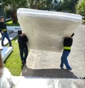 Tres cargadores transportando un gran colchón emplayado al aire libre como parte de unas mudanzas compartidas.