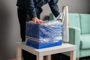 Hombre protegiendo caja azul con plástico film transparente, consejo profesional de como empacar una mudanza sin daños.
