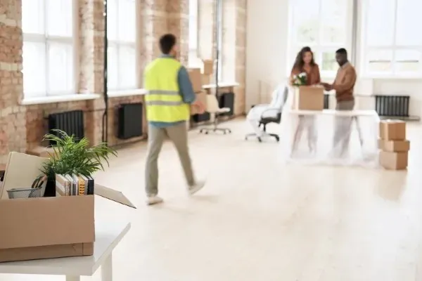 Caja con artículos de escritorio en primer plano y gente trabajando al fondo durante una mudanza de oficina.